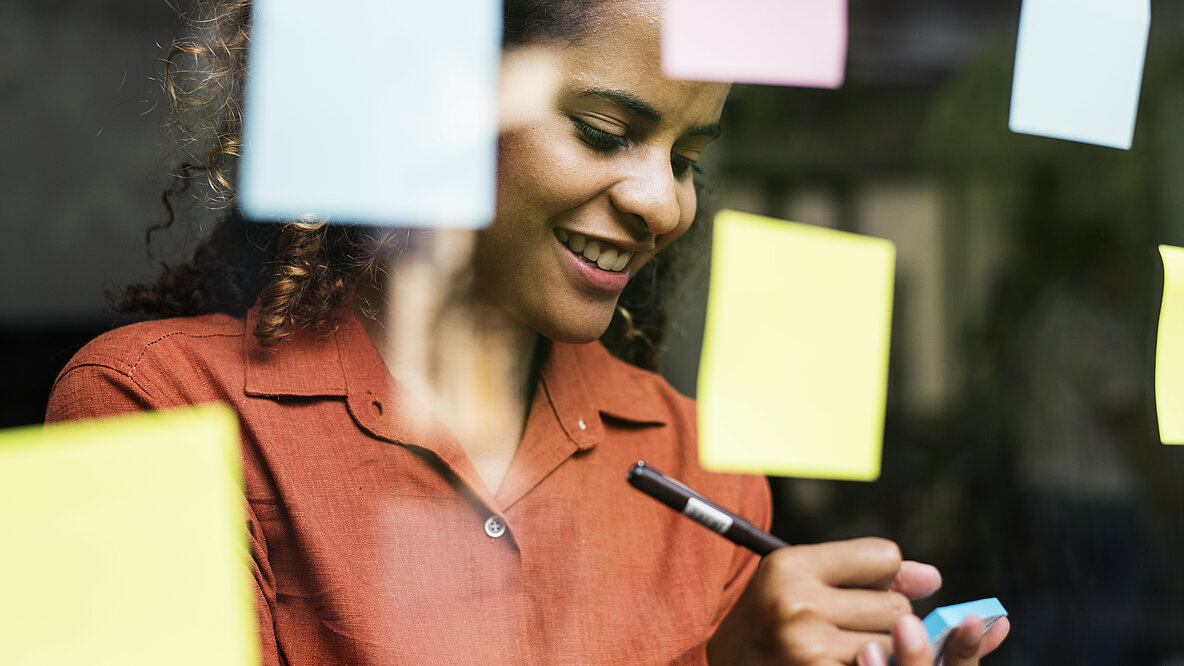 Eine Junge Frau steht vor einer Glasscheibe mit Post-its. Sie macht sich Notizen und lächelt zuversichtlich.