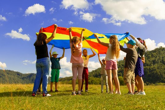 Eine Gruppe Kinder steht auf einer Wiese, sie heben ein buntes Tuch in die Luft, im Hintergrund blauer Himmel und Wolken. 