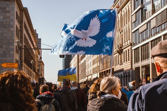 Eine Demonstration in Berlin, viele Personen gehen auf der Straße zwischen zwei Hochhäusern, es wird eine Fahne mit einer Friedenstaube geschwungen sowie eine ukrainische Landesflagge. 