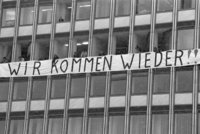 Ein großes Gebäude, aus mehreren Fenster wird ein Transparent am Gebäude gehalten. Auf dem Transparent steht "Wir kommen wieder".