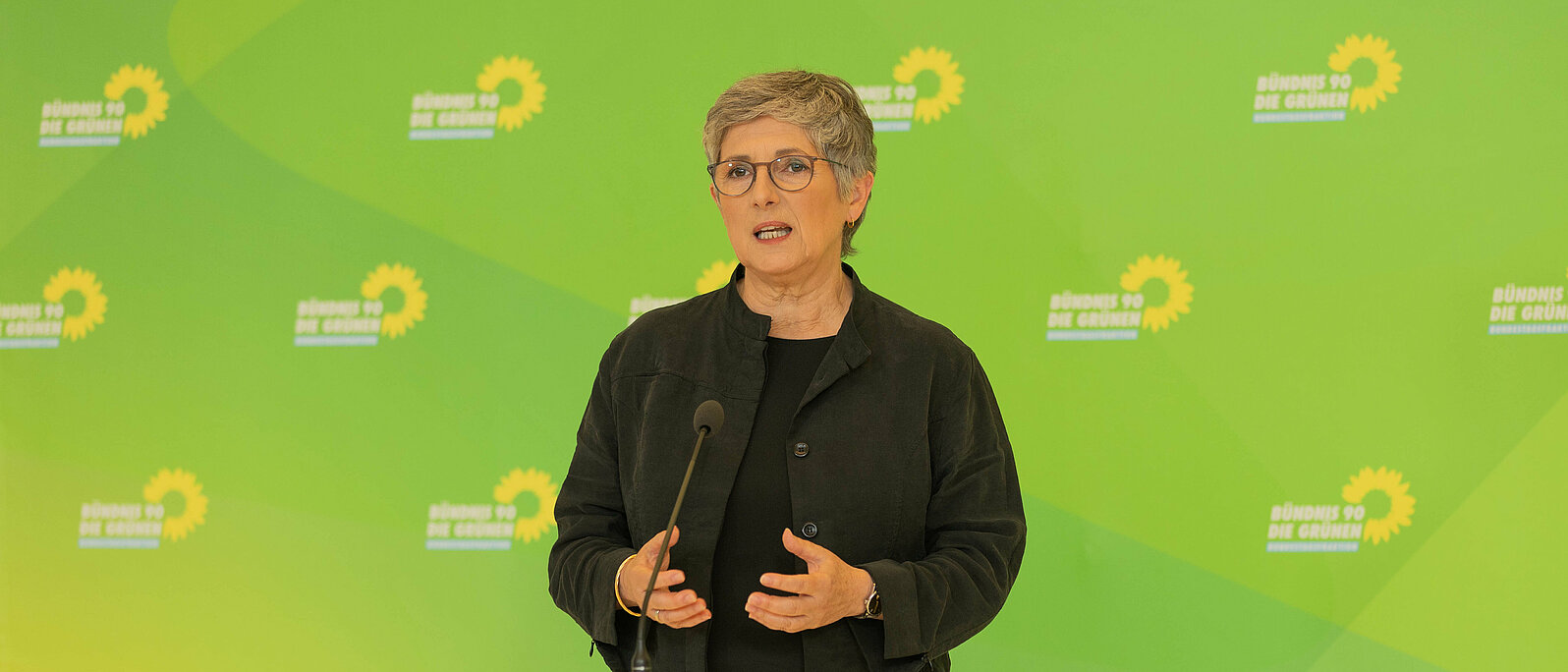 Die Fraktionsvorsitzende Britta Haßelmann steht vor einer grünen Wand und spricht in ein Mikrofon. 
