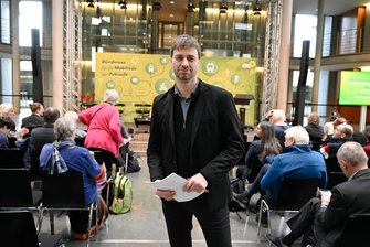 Stefan Gelbhaar steht zwischen den Teilnehmenden der Konferenz. Im Hintergrund ist die Bühne in der Halle des Paul-Löbe-Hauses zu sehen.