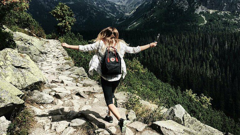 Eine Frau bei der Wanderung im Gebirge: Sie läuft beschwingt über Steine, hebt die Arme und trägt einen Rucksack. Man sieht sie von hinten auf eine schöne Berglandschaft zu laufen.