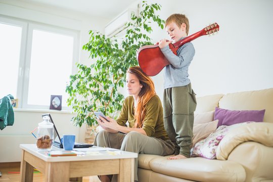 Eine Frau sitzt im Wohnzimmer auf dem Sofa, ein kleiner Junge steht auf dem Sofa und spielt Gitarre.