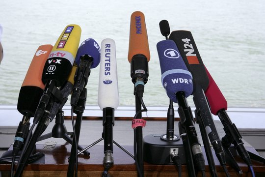 Viele Mikrofone stehen bei einer Pressekonferenz nebeneinander auf einem Tisch. 