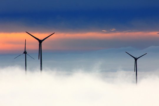 Drei Windkraftanlagen im Nebel, im Hintergrund geht die Sonne auf. 