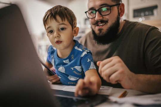 Ein Vater hat seinen Sohn auf dem Schoß, sie sitzen an einem Tisch und schauen auf einen Laptop.