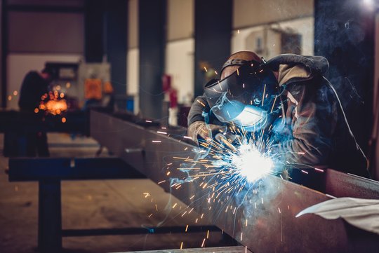 Ein Schweißer in einer industriellen Fertigungshalle, er trägt eine Schutzmaske bei der Arbeit.