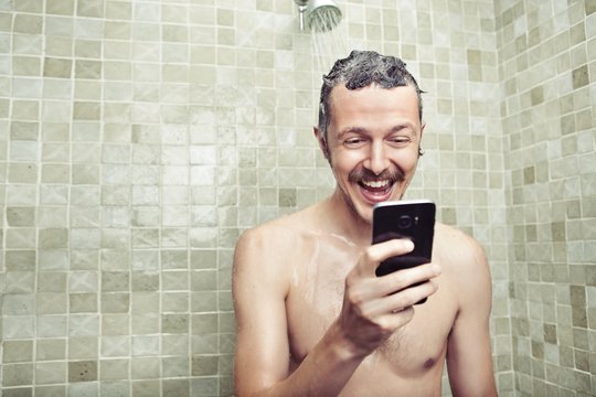 Ein Mann steht unter der Dusche und schaut auf sein Smartphone, er lacht.