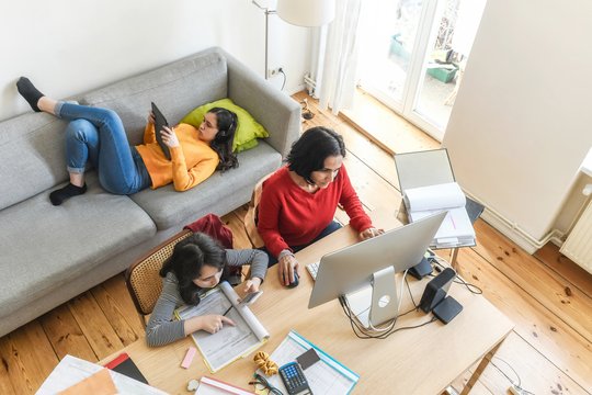 Eine Frau arbeitet am Laptop im Wohnzimmer, zwei Kinder sitzen neben ihr auf Sofa und Stuhl. 