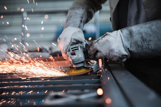 Eine Person bei der Arbeit, ihre Hände werden gezeigt. Die Person trägt Schutzkleidung und schweißt Metallteile zusammen.