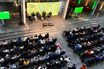 Zu sehen ist die die Bühnensituation in der Halle des Paul-Löbe-Hauses. Auf der Bühne unterhalten sich 5 Personen, hinter Ihnen ist ein großer Bühnenhintergrund mit dem grafischen Veranstaltungsmotiv zu sehen. Vor der Bühne sitzen in 2 Stuhl-Blöcken zahlreiche Teilnehmende.