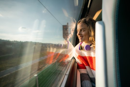 Ein Mädchen sitzt in einem Zug und schaut während der Fahrt lächelnd aus dem Fenster. 