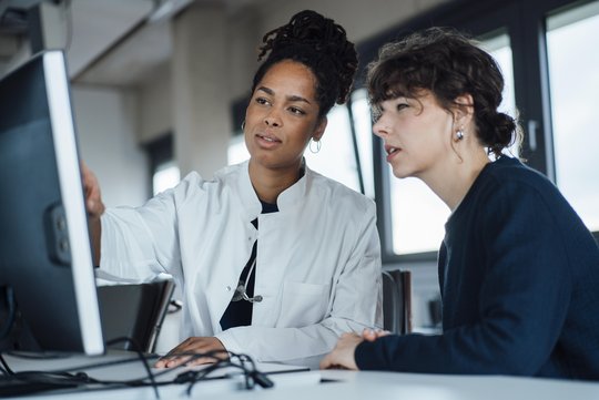 Zwei Frauen sitzen nebeneinander, eine trägt einen Arztkittel. Die Ärztin erklärt der Patientin etwas an einem Computer. 