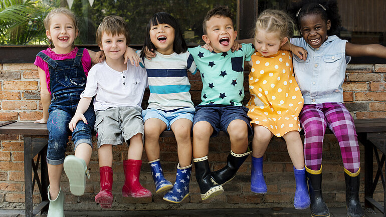 Kita-Kinder sitzen nebeneinander auf einer Bank, sie lachen in die Kamera, alle sind bunt gekleidet und tragen Gummistiefel