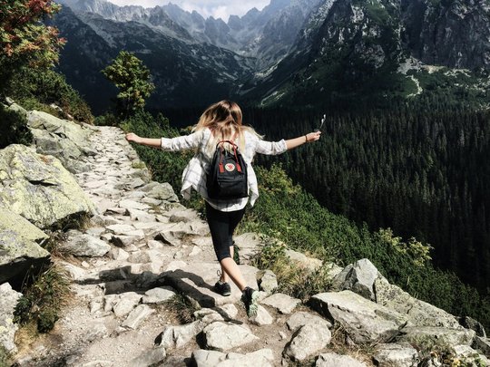 Eine Frau bei der Wanderung im Gebirge: Sie läuft beschwingt über Steine, hebt die Arme und trägt einen Rucksack. Man sieht sie von hinten auf eine schöne Berglandschaft zu laufen.