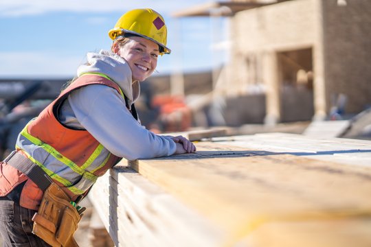 Eine Bauarbeiterin auf einer Baustelle, sie trägt Sicherheitskleidung und lächelt in die Kamera.