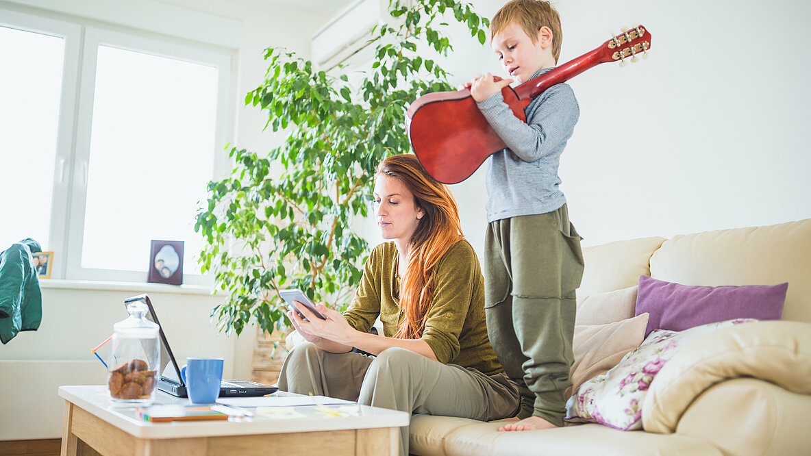 Eine Frau sitzt auf dem Sofa und schaut auf ihr Smartphone, neben ihr auf dem Sofa steht ein kleiner Junge mit einer Gitarre in der Hand.