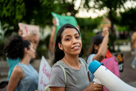 Eine junge Frau bei einer Demonstration, sie hat ein Megafon in der Hand und blickt in die Kamera. Im Hintergrund weitere protestierende Personen.