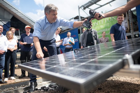 Robert Habeck beugt sich über eine Solaranlage und greift einen Akkubohrschrauber, im Hintergrund stehen Personen.