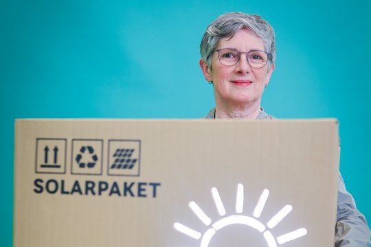 Die Grüne Fraktionsvorsitzende Britta Haßelmann steht mit einem großen Karton vor einer blaugrünen Wand. Auf dem Karton steht Solarpaket