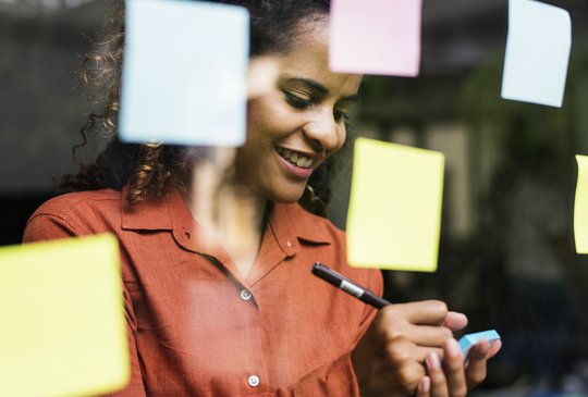 Eine Junge Frau steht vor einer Glasscheibe mit Post-its. Sie macht sich Notizen und lächelt zuversichtlich.