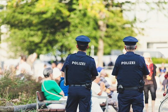 Zwei Polizisten stehen nebeneinander, sie sind von hinten zu sehen und schauen in einen Park, in dem sich Personen aufhalten. 