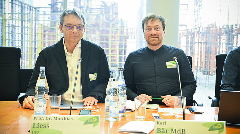 Der Abgeordnete Karl Bär sitzt gemeinsam mit einem weiteren Teilnehmer sitzt an einem Tisch in einem Sitzungssaal.