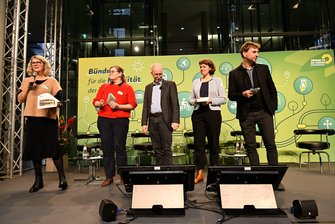 5 Personen stehen auf der Bühne in der Halle, Susanne Menge spricht ins Handmikrofon.