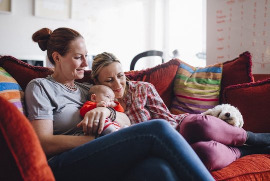 Eine Familie sitzt auf einem Sofa: Ein gleichgeschlechtliches Paar genießt gemeinsames Kuscheln zu Hause, mit einem Baby einem Hund.