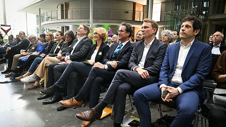 Im Publikum der Veranstaltung sitzen in der ersten Reihe auf Stühlen nebeneinander die Abgeordneten Jürgen Kretz, an-Niclas Gesenhues, Harald Ebner und weitere Gäste.